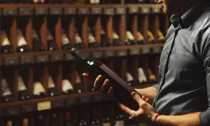 Uomo in cantina esamina una bottiglia di vino, chiedendosi se il vino scade. Scaffali pieni di bottiglie sullo sfondo.
