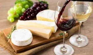 Un bicchiere di vino rosso viene versato, accanto a un bicchiere di vino bianco, con un assortimento di formaggi e uva su un tagliere.