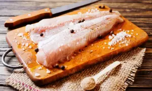 Filetti di nasello pesce pronti per essere cucinati, conditi con sale grosso e pepe su un tagliere di legno.