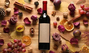Bottiglia di vino scuro con etichetta vuota, circondata da ingredienti autunnali: prugne, uva, arance essiccate, rose secche, cannella, noci e foglie. Parti della bottiglia e del suo contenuto sono visibili.