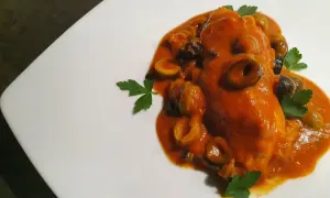 Un saporito pesce scorfano in salsa rossa con olive nere e verdi, guarnito con prezzemolo fresco su un piatto bianco.
