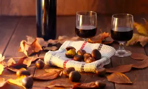 Vino per castagne su un tavolo di legno, circondato da foglie autunnali. Due bicchieri di vino rosso e una ciotola di castagne.