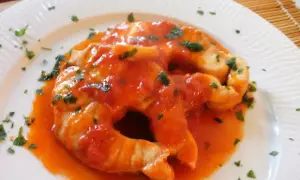 Due fette di pesce palombo cotte in salsa di pomodoro e guarnite con prezzemolo tritato, servite su un piatto bianco.
