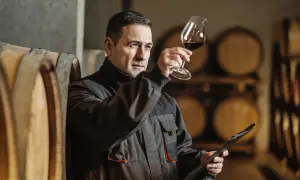 Uomo in cantina osserva il vino in un calice, con botti sullo sfondo. Il suo sguardo attento rivela il **taglio significato** del suo lavoro.