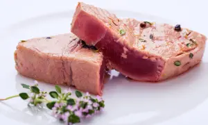 Due tranci di tonno pesce, conditi con erbe e pepe, su un piatto bianco, con un rametto di fiori viola in primo piano.