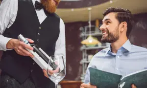 Cameriere con barba presenta una bottiglia di vino a un cliente sorridente che legge il menu. Si chiede a chi si versa prima il vino.
