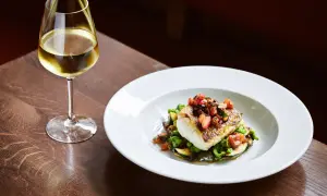 Un bicchiere di vino per pesce, accanto a un piatto di pesce bianco con verdure e pomodoro.
