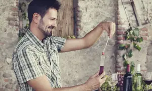 Uomo sorridente misura l'acidità totale del vino rosso con un densimetro. Sullo sfondo, bottiglie di vino e grappoli d'uva.