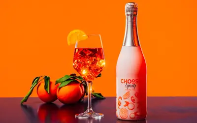 Un bicchiere di spritz ricetta perfetta, con ghiaccio e arancia, accanto a una bottiglia e arance fresche.