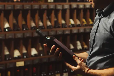 Uomo in cantina esamina una bottiglia di vino, chiedendosi se il vino scade. Scaffali pieni di bottiglie sullo sfondo.