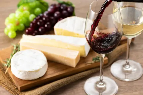 Un bicchiere di vino rosso viene versato, accanto a un bicchiere di vino bianco, con un assortimento di formaggi e uva su un tagliere.