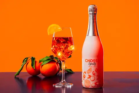 Un bicchiere di spritz ricetta perfetta, con ghiaccio e arancia, accanto a una bottiglia e arance fresche.