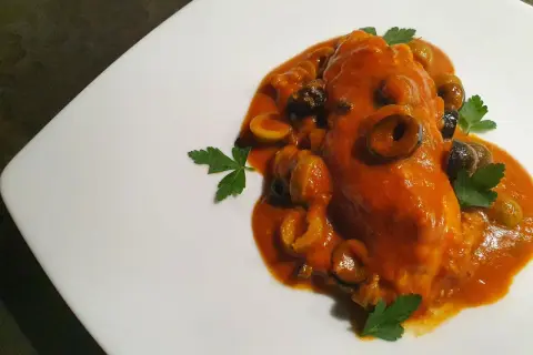 Un saporito pesce scorfano in salsa rossa con olive nere e verdi, guarnito con prezzemolo fresco su un piatto bianco.