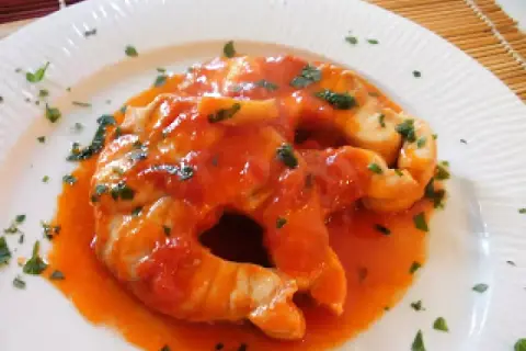 Due fette di pesce palombo cotte in salsa di pomodoro e guarnite con prezzemolo tritato, servite su un piatto bianco.