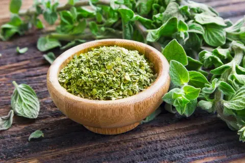 Un ciotola di legno piena di origano secco, con rametti freschi sullo sfondo. Il profumo di origano inebria l'aria.