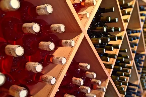 Scaffali di legno pieni di bottiglie di vino rosso, alcune con etichette visibili. Una piccola fioretta di vino attende di essere gustata.