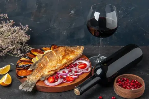 Un gustoso pesce fritto con melanzane, limone e cipolla, accompagnato da un bicchiere e una bottiglia di vino rosso da abbinare al pesce.