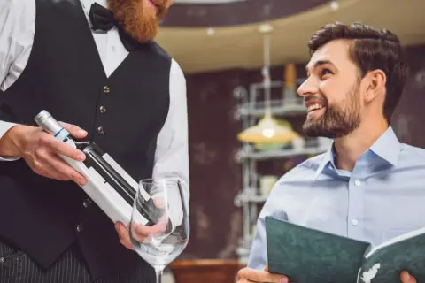 Cameriere con barba presenta una bottiglia di vino a un cliente sorridente che legge il menu. Si chiede a chi si versa prima il vino.