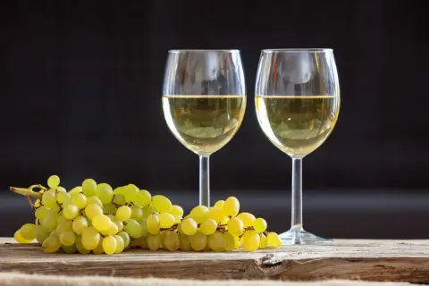 Due calici di vino bianco, tra i migliori vini bianchi italiani, accanto a un grappolo d'uva su un tavolo di legno.