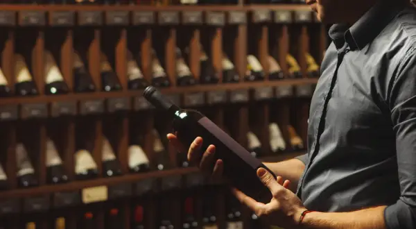 Uomo in cantina esamina una bottiglia di vino, chiedendosi se il vino scade. Scaffali pieni di bottiglie sullo sfondo.