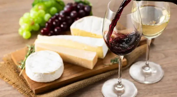 Un bicchiere di vino rosso viene versato, accanto a un bicchiere di vino bianco, con un assortimento di formaggi e uva su un tagliere.