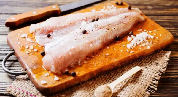 Filetti di nasello pesce pronti per essere cucinati, conditi con sale grosso e pepe su un tagliere di legno.