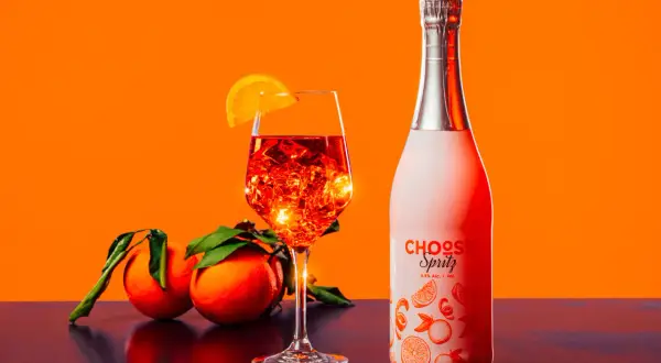 Un bicchiere di spritz ricetta perfetta, con ghiaccio e arancia, accanto a una bottiglia e arance fresche.