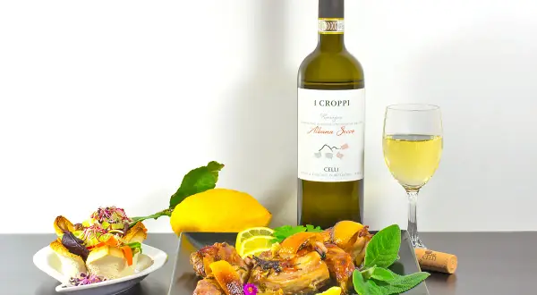 Piatto di coniglio arrosto con limone e erbe aromatiche, accompagnato da un bicchiere e una bottiglia di vino Albana Secco, perfetto vino da abbinare al coniglio.