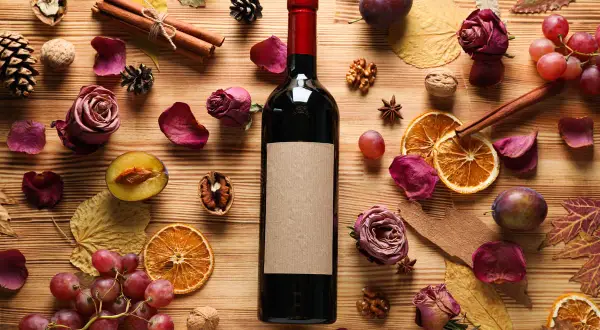 Bottiglia di vino scuro con etichetta vuota, circondata da ingredienti autunnali: prugne, uva, arance essiccate, rose secche, cannella, noci e foglie. Parti della bottiglia e del suo contenuto sono visibili.
