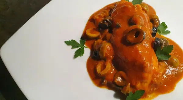 Un saporito pesce scorfano in salsa rossa con olive nere e verdi, guarnito con prezzemolo fresco su un piatto bianco.