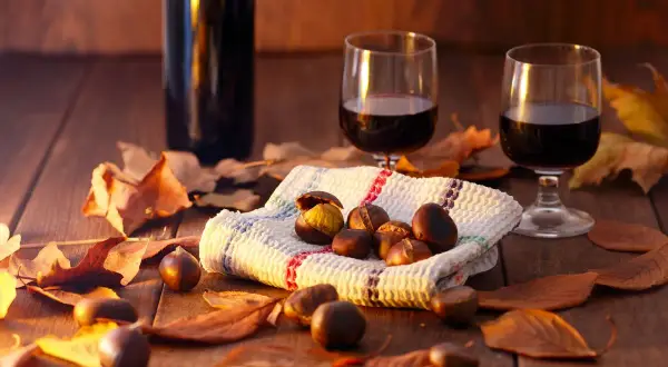 Vino per castagne su un tavolo di legno, circondato da foglie autunnali. Due bicchieri di vino rosso e una ciotola di castagne.