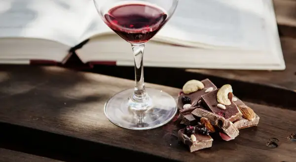 Un bicchiere di vino rosso e cioccolato con frutta secca e noci, un perfetto abbinamento vino e cioccolato, accanto a un libro aperto.