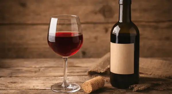 Un bicchiere di vino rosso, una bottiglia con etichetta vuota e un tappo vino su un tavolo di legno rustico.