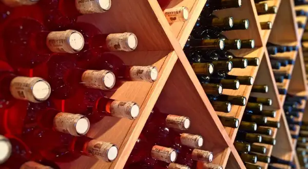 Scaffali di legno pieni di bottiglie di vino rosso, alcune con etichette visibili. Una piccola fioretta di vino attende di essere gustata.