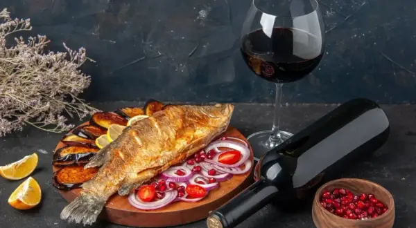 Un gustoso pesce fritto con melanzane, limone e cipolla, accompagnato da un bicchiere e una bottiglia di vino rosso da abbinare al pesce.