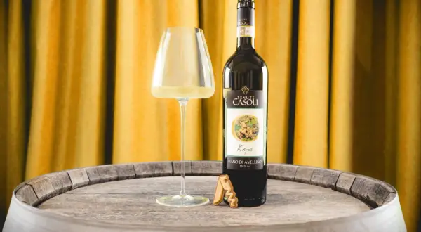 Un bicchiere di Fiano di Avellino DOCG e una bottiglia di vino Fiano di Avellino, su un vecchio barile di legno, con uno sfondo di tende dorate.