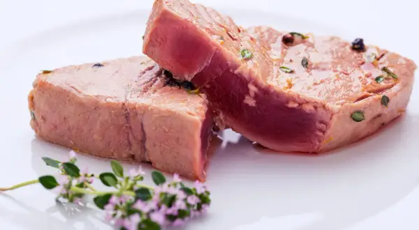 Due tranci di tonno pesce, conditi con erbe e pepe, su un piatto bianco, con un rametto di fiori viola in primo piano.