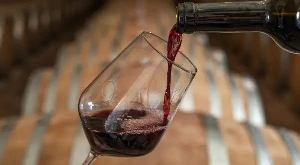 Versando vino rosso in un bicchiere, con botti di legno sullo sfondo. Attenzione, il vino contiene nichel.