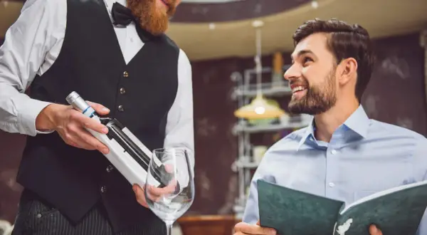 Cameriere con barba presenta una bottiglia di vino a un cliente sorridente che legge il menu. Si chiede a chi si versa prima il vino.