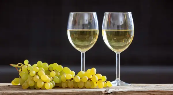 Due calici di vino bianco, tra i migliori vini bianchi italiani, accanto a un grappolo d'uva su un tavolo di legno.