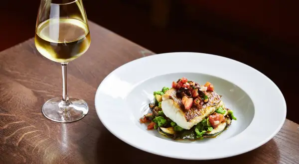 Un bicchiere di vino per pesce, accanto a un piatto di pesce bianco con verdure e pomodoro.