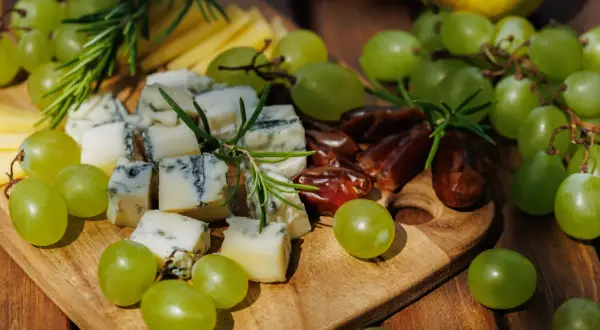 Un tagliere di legno con formaggio erborinato, uva verde succosa, datteri e rosmarino. Un perfetto abbinamento uva e formaggio per un aperitivo.