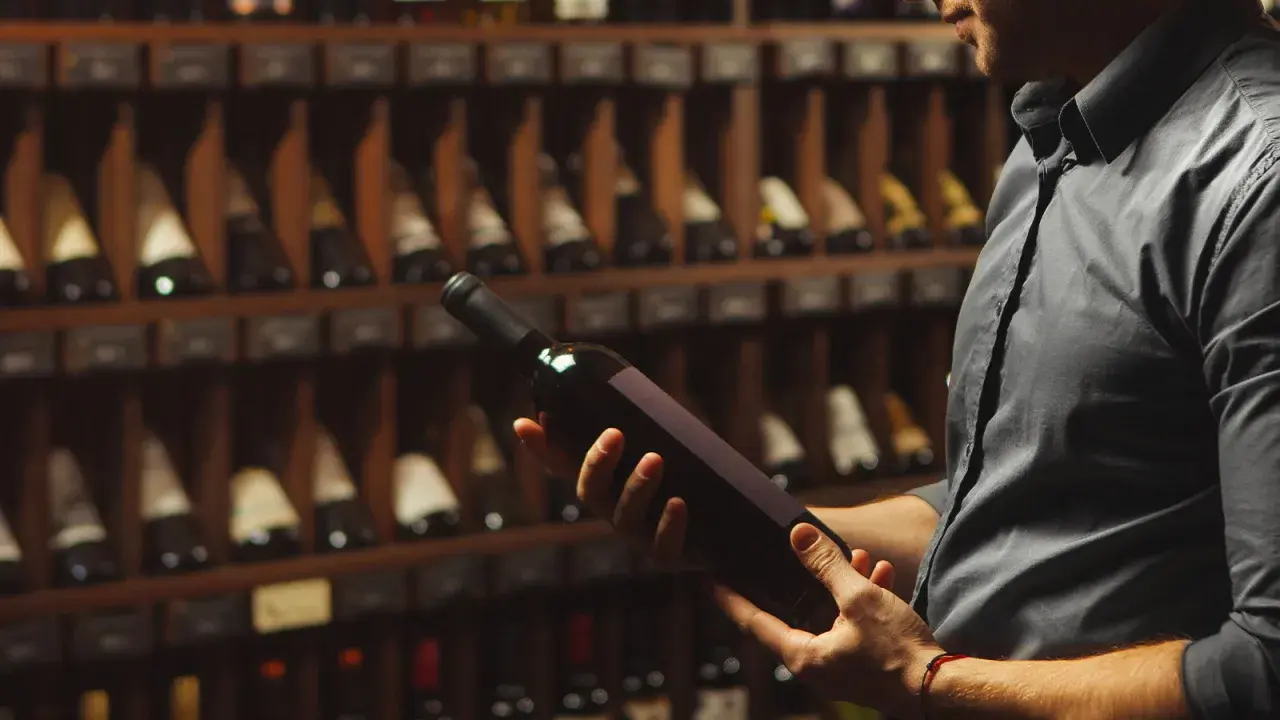 Uomo in cantina esamina una bottiglia di vino, chiedendosi se il vino scade. Scaffali pieni di bottiglie sullo sfondo.