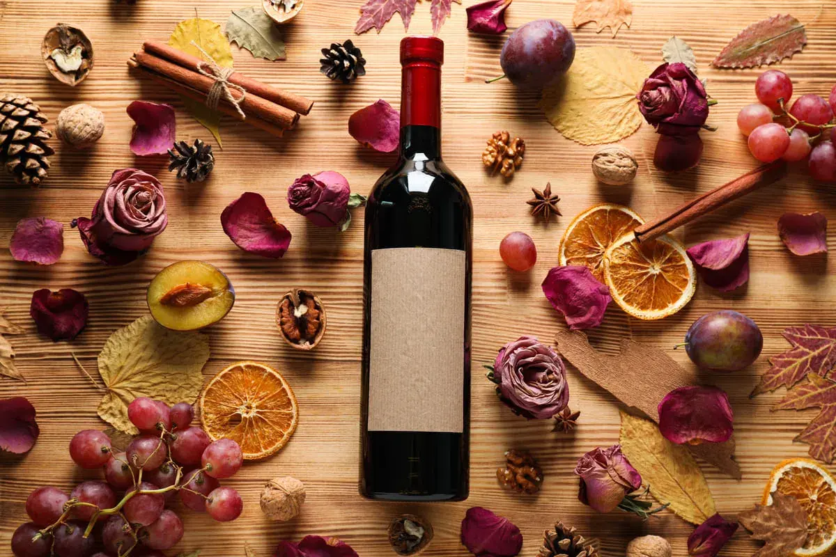Bottiglia di vino scuro con etichetta vuota, circondata da ingredienti autunnali: prugne, uva, arance essiccate, rose secche, cannella, noci e foglie. Parti della bottiglia e del suo contenuto sono visibili.