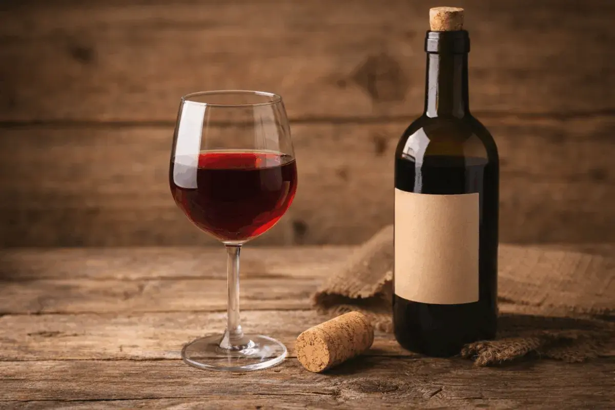 Un bicchiere di vino rosso, una bottiglia con etichetta vuota e un tappo vino su un tavolo di legno rustico.