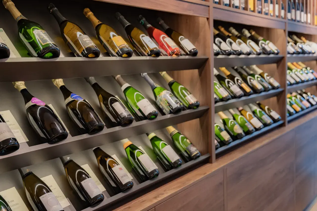 Scaffali pieni di bottiglie di champagne, alcune con etichette dorate e verdi. Una bottiglia da 5 litri nome è visibile in primo piano.