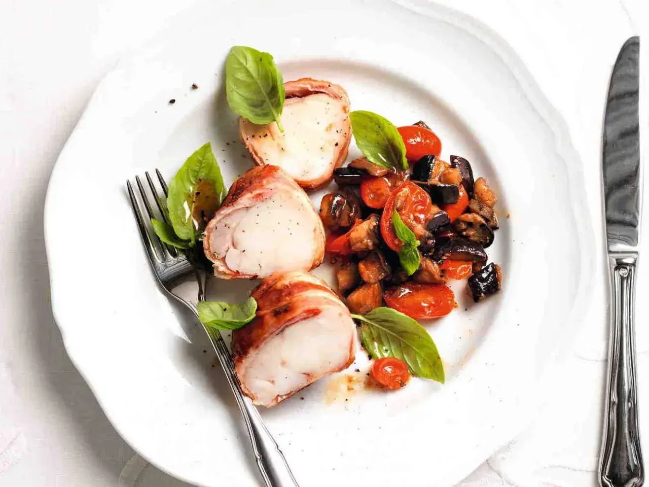 Tranci di rana pescatrice avvolti nel prosciutto, serviti con ratatouille di verdure e basilico fresco.