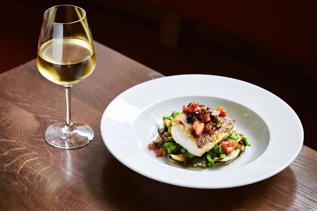 Un bicchiere di vino per pesce, accanto a un piatto di pesce bianco con verdure e pomodoro.