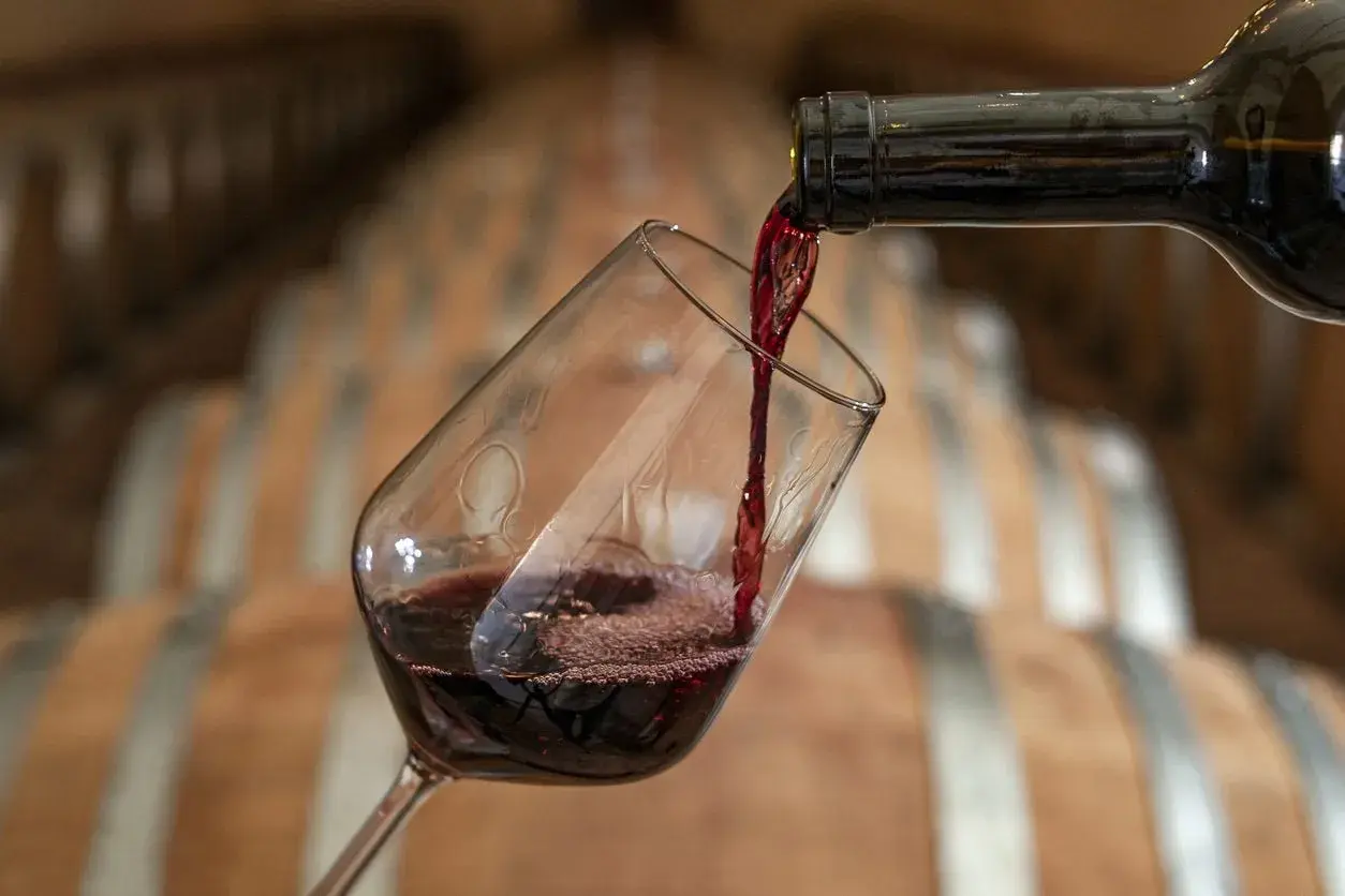 Versando vino rosso in un calice, con botti di legno sullo sfondo. Un calice di vino quanti ml? La risposta è nella degustazione.