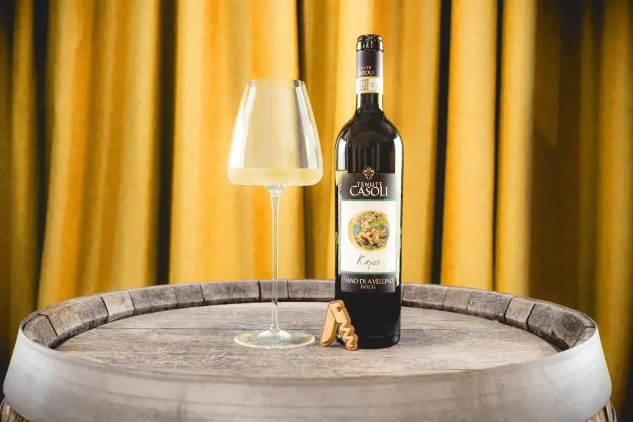 Un bicchiere di Fiano di Avellino DOCG e una bottiglia di vino Fiano di Avellino, su un vecchio barile di legno, con uno sfondo di tende dorate.